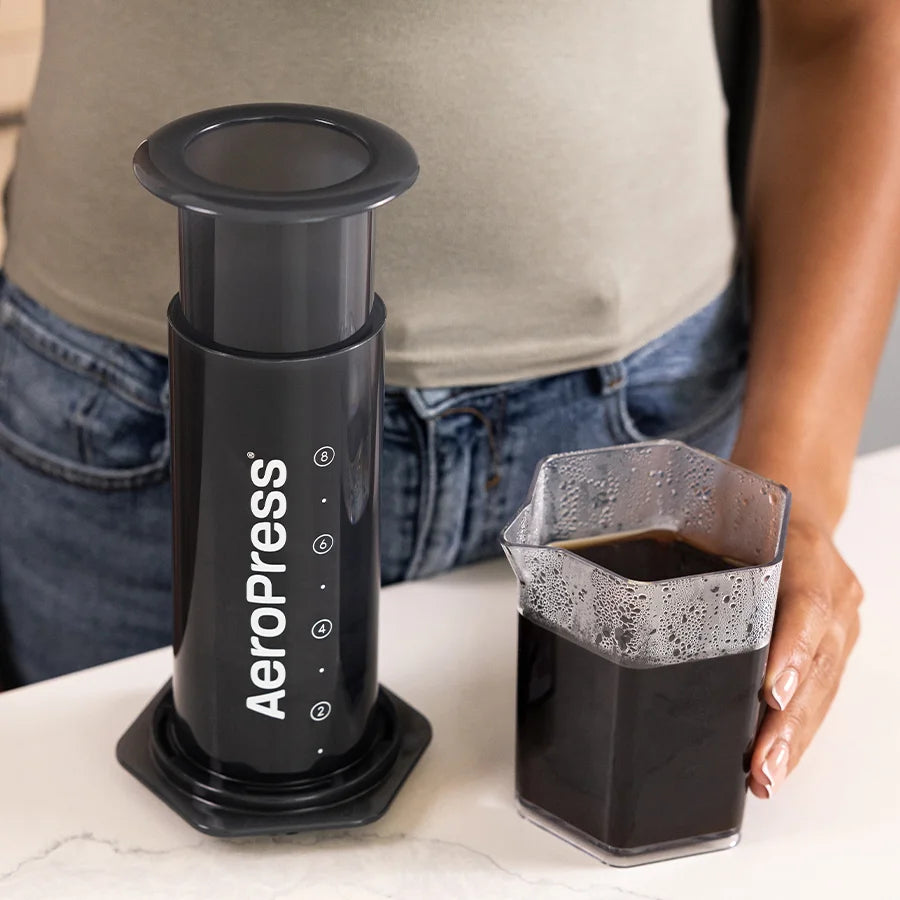 Prensa de café negra con taza de café y mano sosteniendo el vaso