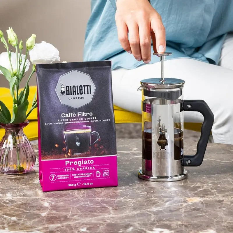 Bolsa de café molido Bialetti Pregiato y cafetera de acero con mano sobre el émbolo