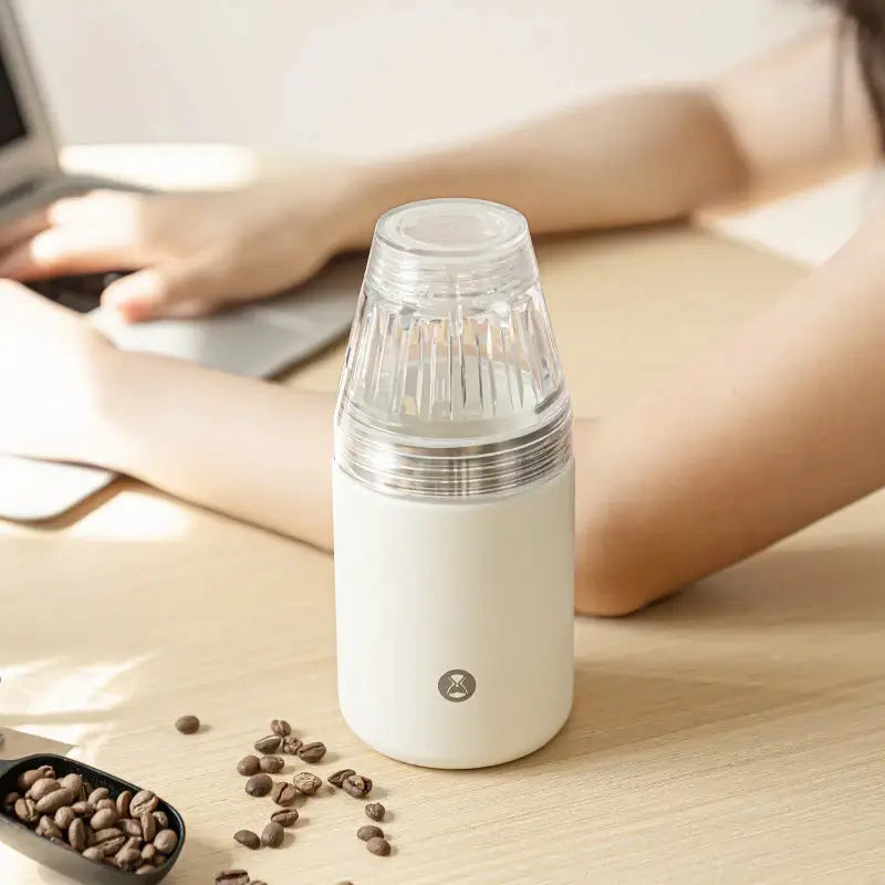 Cafetera portátil blanca con filtro de vidrio cónico, sobre mesa con granos