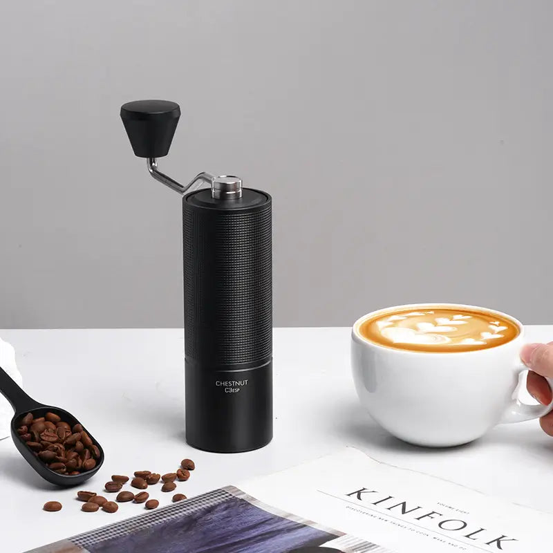 Molino de café negro alto con manivela; junto a granos y taza de café