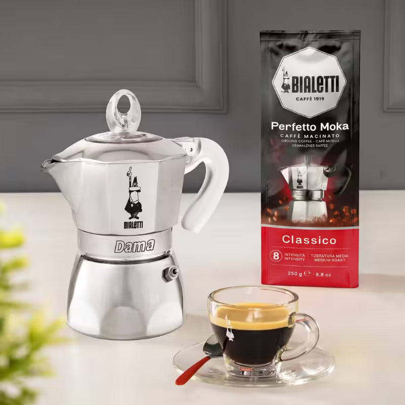 Cafetera Bialetti Dama de acero con taza de café al frente y paquete