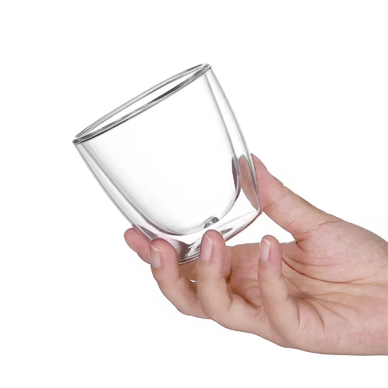 Vaso de vidrio de doble pared transparente sostenido por una mano, vista inclinada